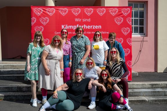 Gruppe von Menschen mit verschiedenen Outfits und Sonnenbrillen vor rotem Hintergrund.