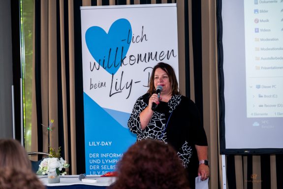 Frau spricht am Mikrofon vor einem Willkommensbanner bei einer Veranstaltung.