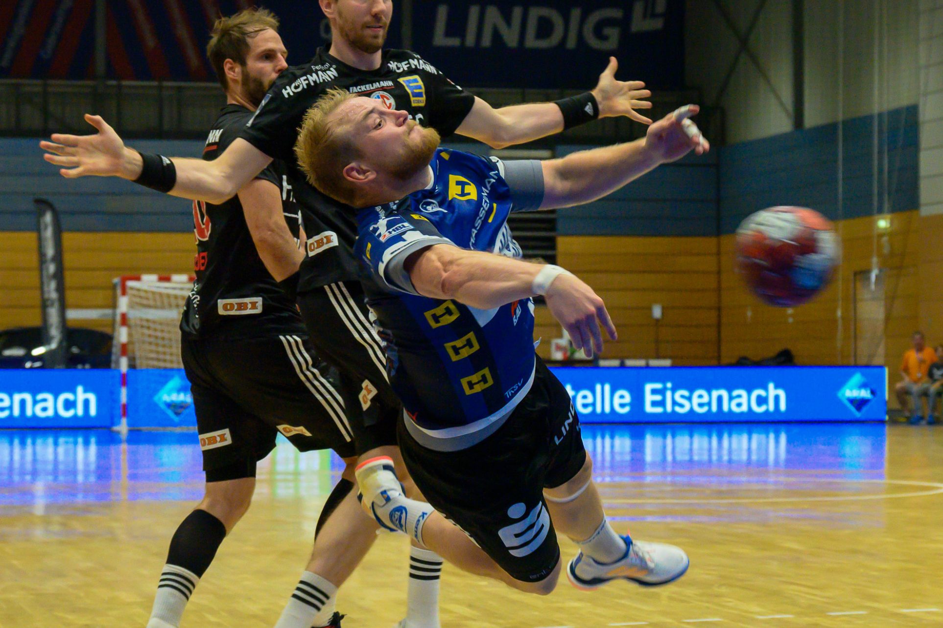 Handballspieler springt zum Wurf, während zwei Verteidiger ihn angreifen.