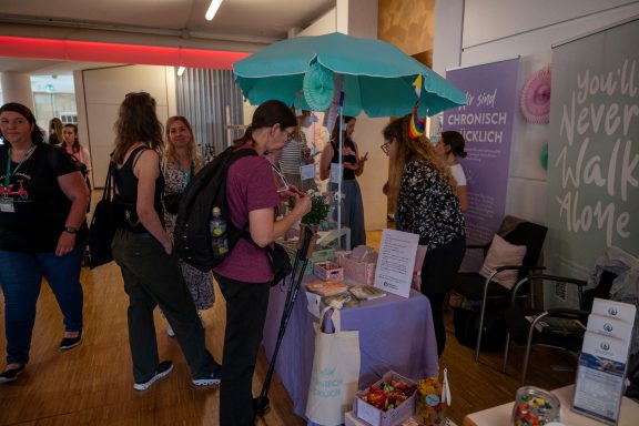 Marktstand mit buntem Sonnenschirm und mehreren Personen, die sich um Produkte versammeln.