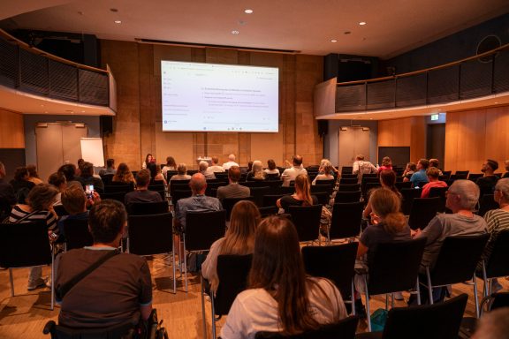Ein Publikum sitzt in einem Veranstaltungsraum, der eine Präsentation verfolgt.