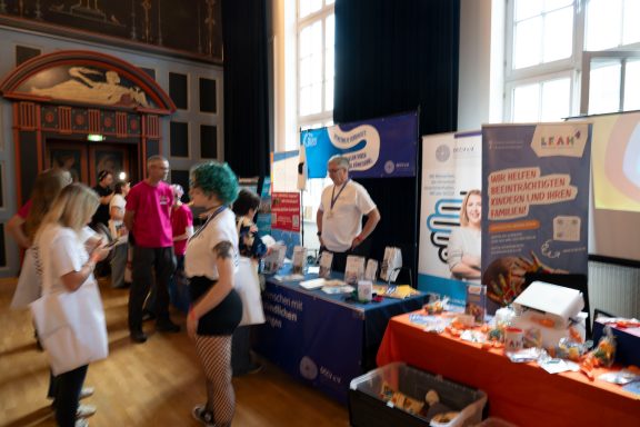 Menschen an Informationsständen in einem Veranstaltungsraum, verschiedene Flyer und Materialien sichtbar.
