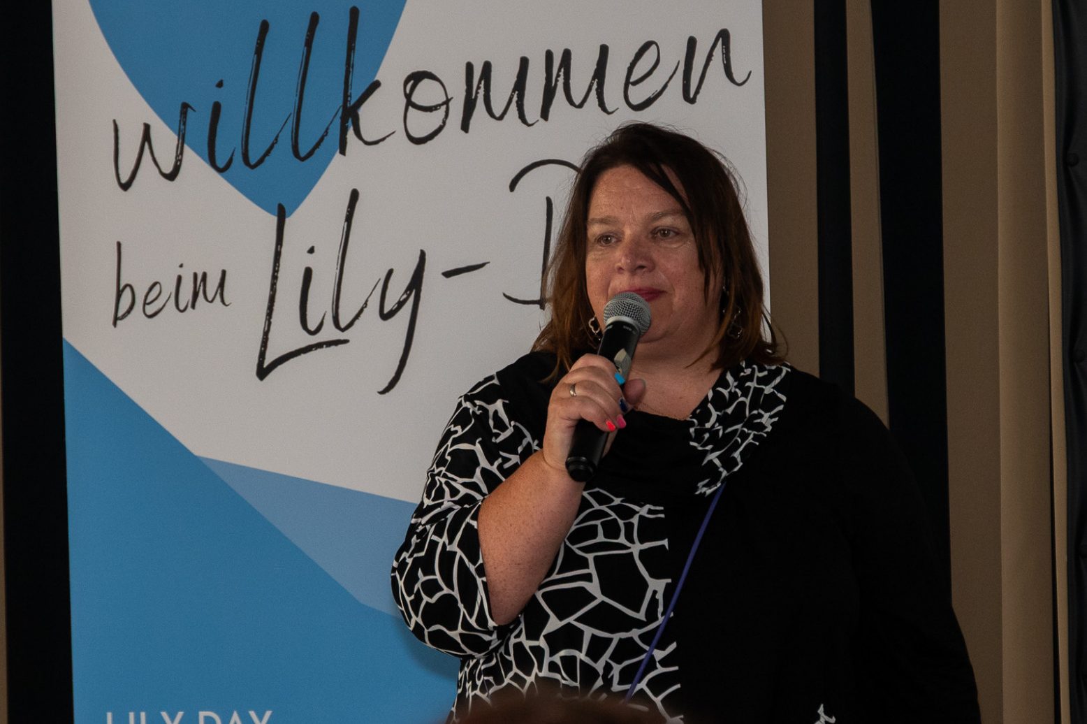 Frau spricht auf einer Veranstaltung, mit einem Mikrofon in der Hand, im Hintergrund ein Banner.