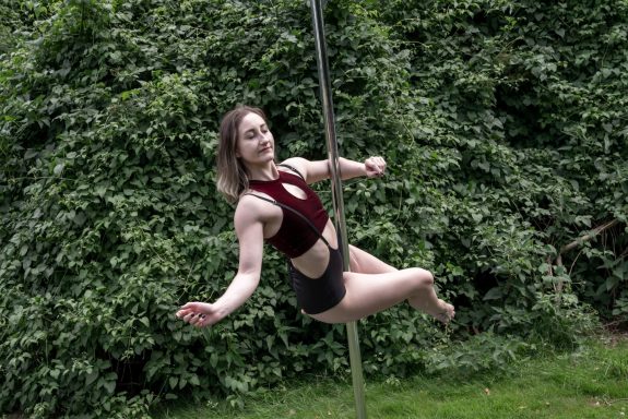 Junge Frau, die an einer Pole-Tanzstange schwingt, umgeben von grünen Bäumen.