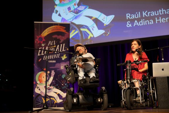 Zwei Personen auf der Bühne, eine im Rollstuhl, die andere sitzt, vor einem Banner.